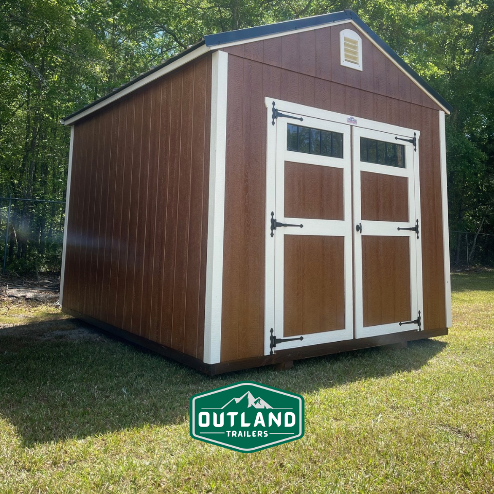 Outland Trailers 10x12 utility shed with metal walls and a peaked roof, set on a grassy area with a door and window.