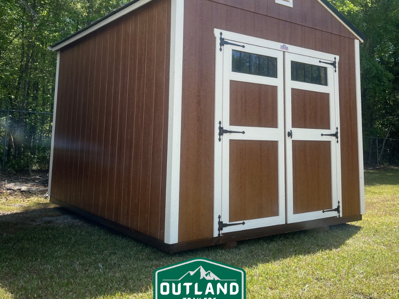Outland Trailers 10x12 utility shed with metal walls and a peaked roof, set on a grassy area with a door and window.