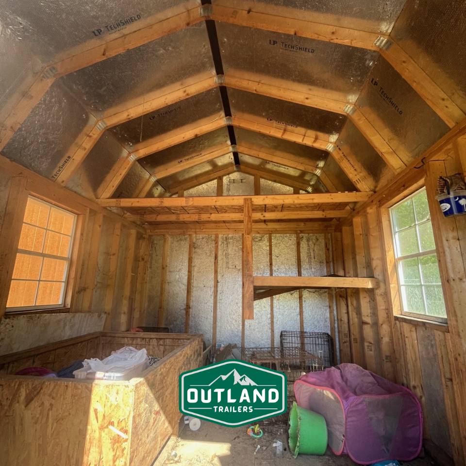 Used Outland Trailers 10x12 lofted barn with raised roof, wooden siding, and double doors, set on a grassy field.