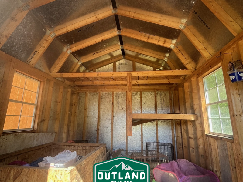 Used Outland Trailers 10x12 lofted barn with raised roof, wooden siding, and double doors, set on a grassy field.